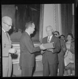 Fotografia "Entrega de Espadim ao representante do embaixador dos EUA. Sr. Mean Gordon representante do embaixador Linconl Gordon, recebe das mães do Cel. Gustavo Borges, espadim oferecido pelo Cel. Edson Moura Freitas, comandante da Polícia Militar, Reportagem de Nelson Silva" ([Local n/d] , 1963) [negativo]. / Fotógrafo(a): Méra.  -- ITEM-0005.