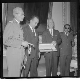 Fotografia "Entrega de Espadim ao representante do embaixador dos EUA. Sr. Mean Gordon representante do embaixador Linconl Gordon, recebe das mães do Cel. Gustavo Borges, espadim oferecido pelo Cel. Edson Moura Freitas, comandante da Polícia Militar, Reportagem de Nelson Silva" ([Local n/d] , 1963) [negativo]. / Fotógrafo(a): Méra.  -- ITEM-0003.